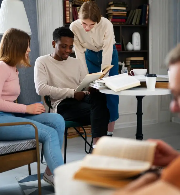 A group of young people sitting together in a cozy room, reading books and discussing them around a small table.